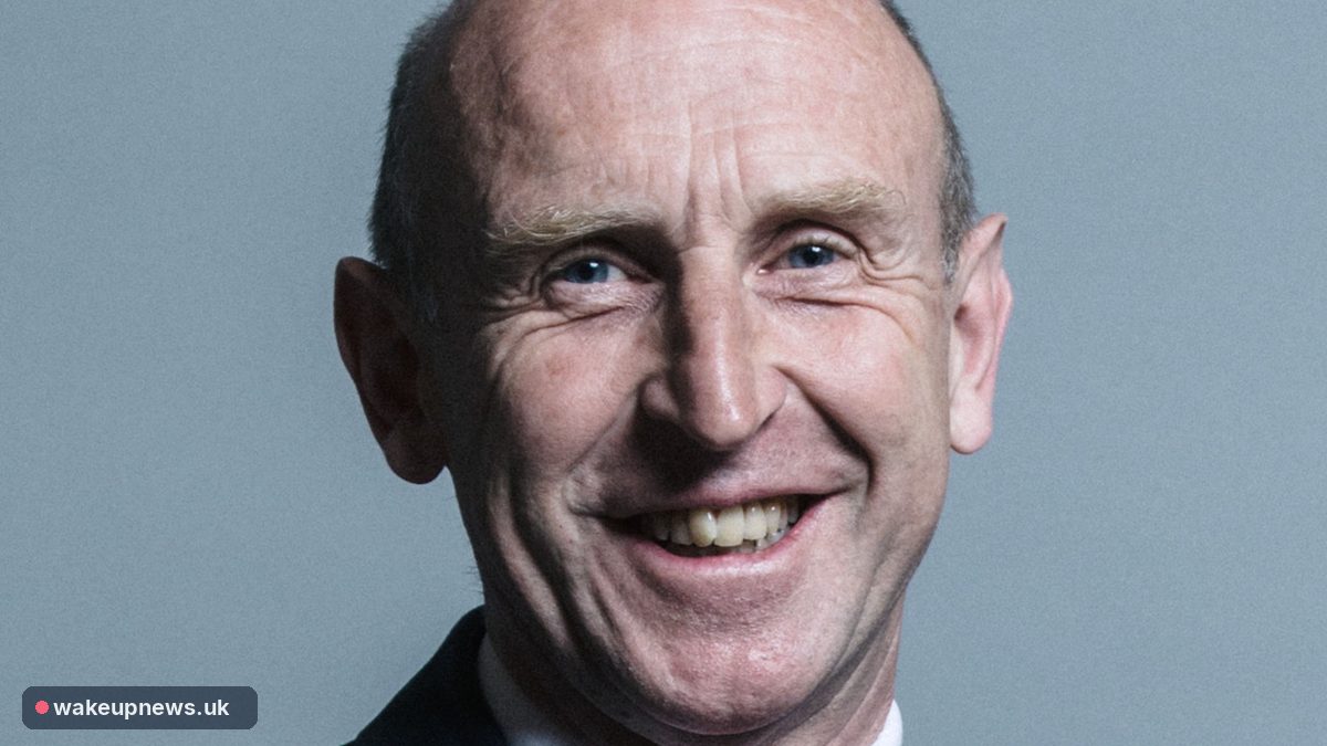 John Healey smiling in an official portrait against a grey-blue background