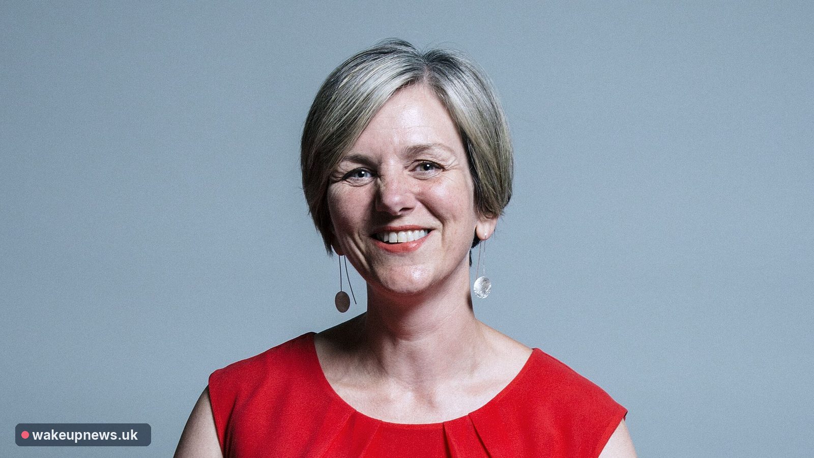 Lilian Greenwood MP smiling, wearing a red top, against a plain blue-grey background